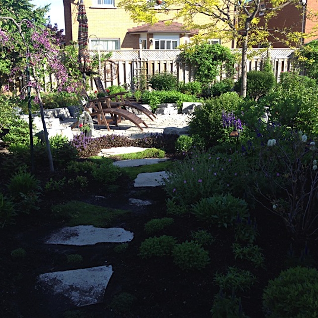 A garden with a stone pathway and chairs in it with stone borders with bushy plants.