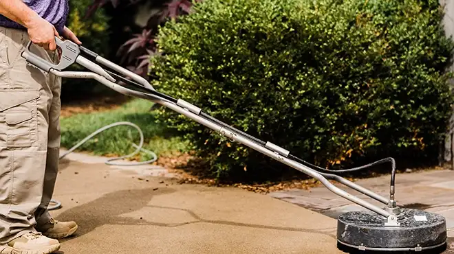 A stainless steel pressure washer surface cleaner being used on a sidewalk, with a bush and lawn in the background.