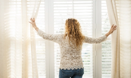 Woman opening curtains on a big, bright window with blinds.