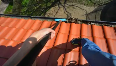 Person on an orange tile roof using a telescope brush to clean the gutter.