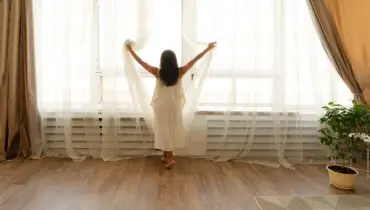 A young girl in white gown opening curtains in front of a large window letting in the morning sunlight.